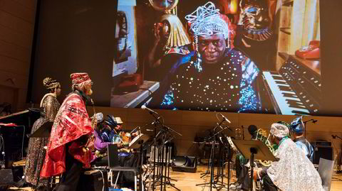 Den snart 100 år gamle Marshall Allen (i rødt) spiller altsaksofon med Sun Ra Arkestra i 2018, under veiledning av avdøde Sun Ra på skjermen.