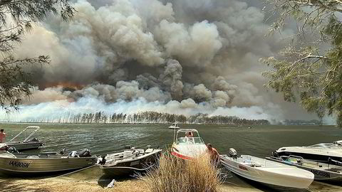 Brannrøyk dekker himmelen over småbåter som er trukket på land ved Lake Conjola i Australia torsdag. Turister evakueres fra utsatte steder sørøst i landet. Foto: Robert Oerlemans / AP / NTB scanpix